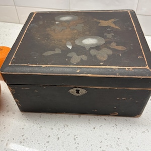 Può includere: Una scatola di legno nera con un disegno floreale e un uccello sul coperchio. La scatola ha un buco della serratura ed è appoggiata su una superficie bianca. Due arance sono visibili sul lato sinistro della scatola.