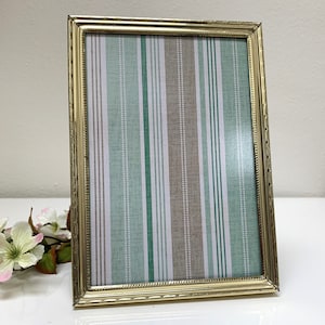 May include: A rectangular gold-colored picture frame with a striped pattern in shades of green, white, and brown. The frame is standing upright on a white surface, with a small floral arrangement in the lower left corner.