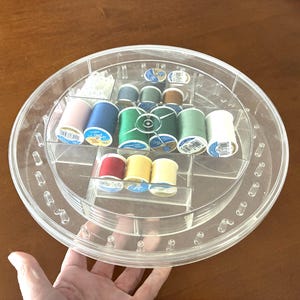 Może przedstawiać: Przezroczysty plastikowy organizer do szycia wypełniony szpulkami nici w różnych kolorach, w tym różowym, niebieskim, zielonym, czerwonym i żółtym. Organizer jest okrągły z wieloma przegródkami i obrotową konstrukcją. Nici są starannie ułożone.