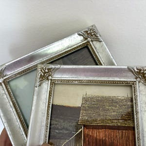 May include: Two rectangular picture frames with a metallic silver finish and ornate corner detailing. The frames feature a beaded inner border. One frame contains a print of a red barn in a landscape, while the other holds a blank light-colored insert.