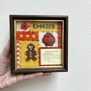 May include: A square needlepoint wall art piece in a dark wood frame featuring a yellow background with a gingerbread man, a red cookie jar, and the text COOKIES. A white card displays a recipe for GINGERBREAD COOKIES with instructions to bake at 375°F.