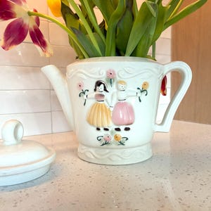May include: White ceramic teapot-shaped vase with a lid, featuring a decorative relief of two figures in dresses. The vase holds fresh tulips. The lid is oval-shaped with a small handle. The teapot has a spout and a handle.
