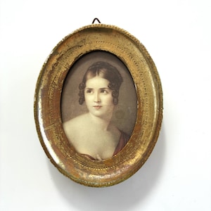 May include: An oval portrait of a woman in a gold-colored frame. The portrait features a woman with dark hair and a red garment. The frame has a distressed, antique appearance, with visible wear and tear.
