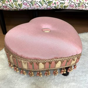 Puede incluir: Un otomán rosa en forma de corazón con una parte superior abotonada. La base está adornada con tela dorada y a rayas, y adornada con borlas colgantes. El otomán se asienta sobre patas cortas y oscuras y está colocado sobre una alfombra blanca.