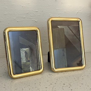 Puede incluir: Dos marcos de fotos rectangulares con bordes de metal dorado pulido y esquinas redondeadas. Cada marco presenta una orientación vertical y un panel de vidrio oscuro que refleja una escena exterior de un edificio. Los marcos se mantienen en pie.