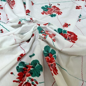 May include: White fabric with a floral pattern featuring red flowers and green leaves. The design includes red and green lines, creating a grid-like structure. The fabric is draped, showing folds and texture.