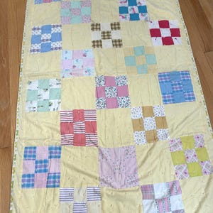 May include: A rectangular patchwork quilt featuring a pale yellow background with scattered tufted knots. The quilt is composed of various square blocks in diverse patterns and colors, including blue, pink, green, red, and brown, finished with a floral border.