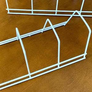 May include: Two white wire organizers, likely for kitchen or office use, are shown on a brown surface. The organizers have a rectangular base with vertical dividers and angled supports, creating a space for storing items. The wire is painted white.