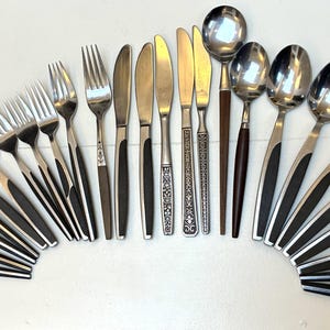 Puede incluir: Una colección de cubiertos de acero inoxidable vintage, que incluye tenedores, cuchillos y cucharas, dispuestos en semicírculo. Los utensilios presentan mangos de color marrón oscuro o negro y una superficie pulida y reflectante.