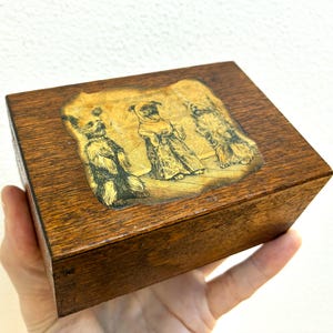 Könnte beinhalten: Kleine, rechteckige Holzbox mit Scharnierdeckel. Die Oberseite der Box zeigt ein goldfarbenes Bild von drei Hunden. Die Box hat eine warme braune Farbe und scheint Vintage zu sein.