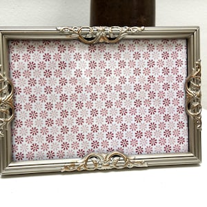 May include: Silver rectangular picture frame with ornate gold accents. The frame holds a patterned insert with a repeating floral design in shades of pink and white. The frame is displayed on a white surface.