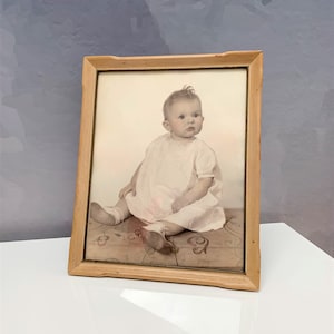 May include: A vintage-style rectangular wooden picture frame containing a sepia-toned portrait of a seated infant wearing a light-colored dress and matching shoes. The frame has a natural light wood finish with a simple, clean-lined border design.