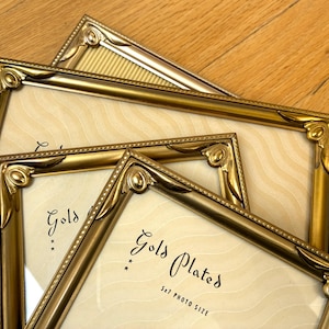 May include: Gold-plated photo frames, 5x7 inches, arranged on a wooden surface. The frames have ornate, decorative corners and a gold finish. The text "Gold Plated" is visible on the cream-colored backing.