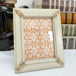 May include: A rectangular, off-white picture frame with decorative corner accents. The frame holds a patterned orange and white print. The frame has a distressed finish, suggesting a vintage or antique style. The overall aesthetic is classic and decorative.