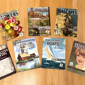 May include: A collection of vintage art instruction books, including titles like "How to Draw and Paint Flowers," "Oil Painting," and "Perspective Drawing." The books feature colorful illustrations and are arranged on a wooden surface.