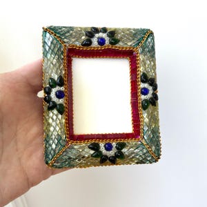 Könnte beinhalten: Rechteckiger Bilderrahmen mit Mosaikdesign. Der Rahmen hat einen roten Innenrand, eine grün-goldene Mosaikaußenseite und ist mit dekorativen floralen Akzenten in Blau, Schwarz und Grün verziert. Eine goldene Kettenverzierung vervollständigt das Design.