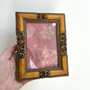 May include: A rectangular picture frame with a mustard yellow finish and dark brown accents. The frame features intricate, carved scrollwork details at the centre of each side. Inside, a red fabric insert displays a gold floral pattern.