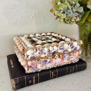 May include: A rectangular wooden box covered in a mosaic of various seashells in shades of white, pink, and purple. The lid and sides are densely decorated with shells of different shapes and sizes, featuring a small gold-toned metal latch closure.