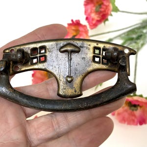 May include: Antique brass drawer pull with a decorative design. The pull features a curved handle and a central design with an arrow-like shape and a heart. The rectangular cutouts add detail to the piece. The hardware is held in a hand.