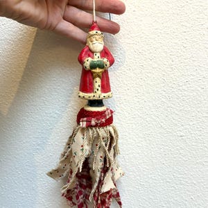 May include: A Christmas ornament featuring a Santa Claus figure in a red coat with a white beard, holding a green gift. Below the figure is a fabric tassel with red and cream-colored fabric strips. The ornament is suspended by a string.