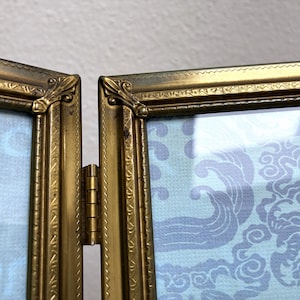 May include: A vintage-style, hinged picture frame with a gold finish. The frame is divided into two sections, connected by a brass hinge. The interior displays a light blue patterned design. The frame's ornate detailing adds to its antique aesthetic.