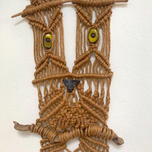 May include: A brown macrame wall hanging featuring a stylized owl face design. The piece is constructed with knotted cord, two yellow ceramic eye beads with dark centers, a dark triangular nose, and two horizontal wooden branches.