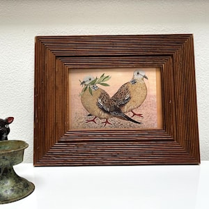 May include: A framed print featuring two doves with olive branches in their beaks. The birds are depicted in shades of brown and tan, set against a soft, pinkish background. The frame is made of dark brown wood with a ridged design.