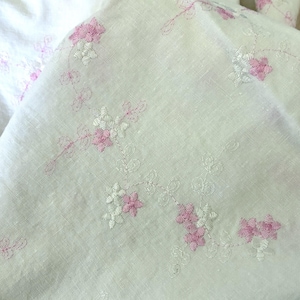 Könnte beinhalten: Off-white Leinenstoff mit gestickten Blumenmustern in Rosa und Weiß. Die Stickerei zeigt zarte Blumen und Ranken, die ein weiches, strukturiertes Design bilden. Der Stoff ist gefaltet und zeigt die detaillierten Nähte.