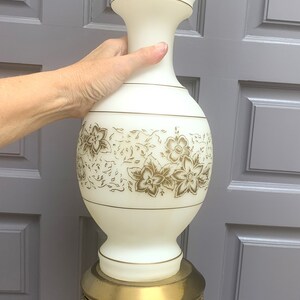 May include: A vintage table lamp with a white, vase-shaped body adorned with gold floral patterns. The lamp features a brass base and accents, and a pull chain. The lamp is approximately 24 inches tall.