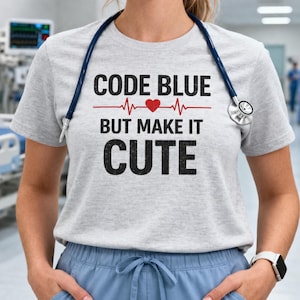 May include: A light gray t-shirt with the text "CODE BLUE BUT MAKE IT CUTE" in black, red, and white. The design includes a heartbeat line and a red heart. A stethoscope is around the neck. The person is wearing blue scrubs.