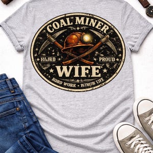 May include: A light grey t-shirt with a distressed graphic reading "COAL MINER WIFE" in a circular design. The design includes a miner's helmet, crossed pickaxes, and the phrases "HARD" and "PROUD". Blue jeans and grey trainers are also visible.