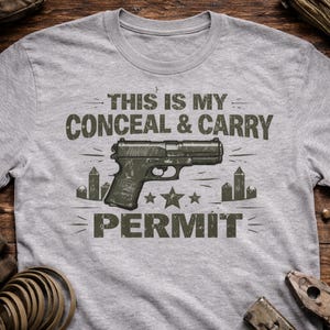 May include: A light grey t-shirt featuring the text "THIS IS MY CONCEAL & CARRY PERMIT" in distressed green lettering. A handgun graphic is centered, with stars and stylized buildings.