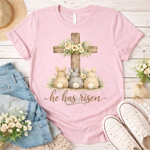 Op de afbeelding: Roze T-shirt met een Paasontwerp. De afbeelding toont drie konijntjes, een houten kruis versierd met bloemen en de tekst "he has risen". Het shirt wordt gecombineerd met een blauwe jeans en witte sneakers.