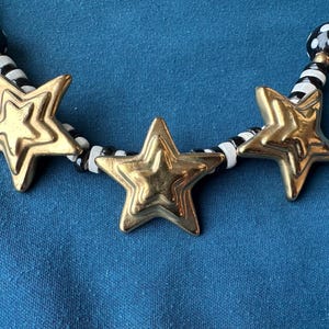 May include: Gold-toned star pendant necklace with black and white beaded accents. The necklace features three layered star pendants and a mix of black, white, and gold beads. The necklace is displayed on a blue background.