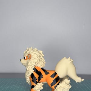 May include: A detailed figurine of an Arcanine, a canine-like Pok&eacute;mon. The figure features an orange body with black stripes, cream-colored mane and tail, and orange paws. The figure is standing on a green grid surface.