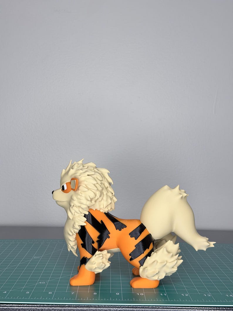 May include: A detailed figurine of an Arcanine, a canine-like Pok&eacute;mon. The figure features an orange body with black stripes, cream-colored mane and tail, and orange paws. The figure is standing on a green grid surface.