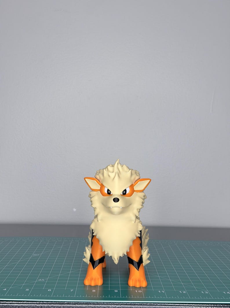 May include: A cream-colored, stylized Arcanine figure with orange and black accents. The figure has a fluffy mane, orange ears, and orange and black markings on its legs. It stands on a green cutting mat.
