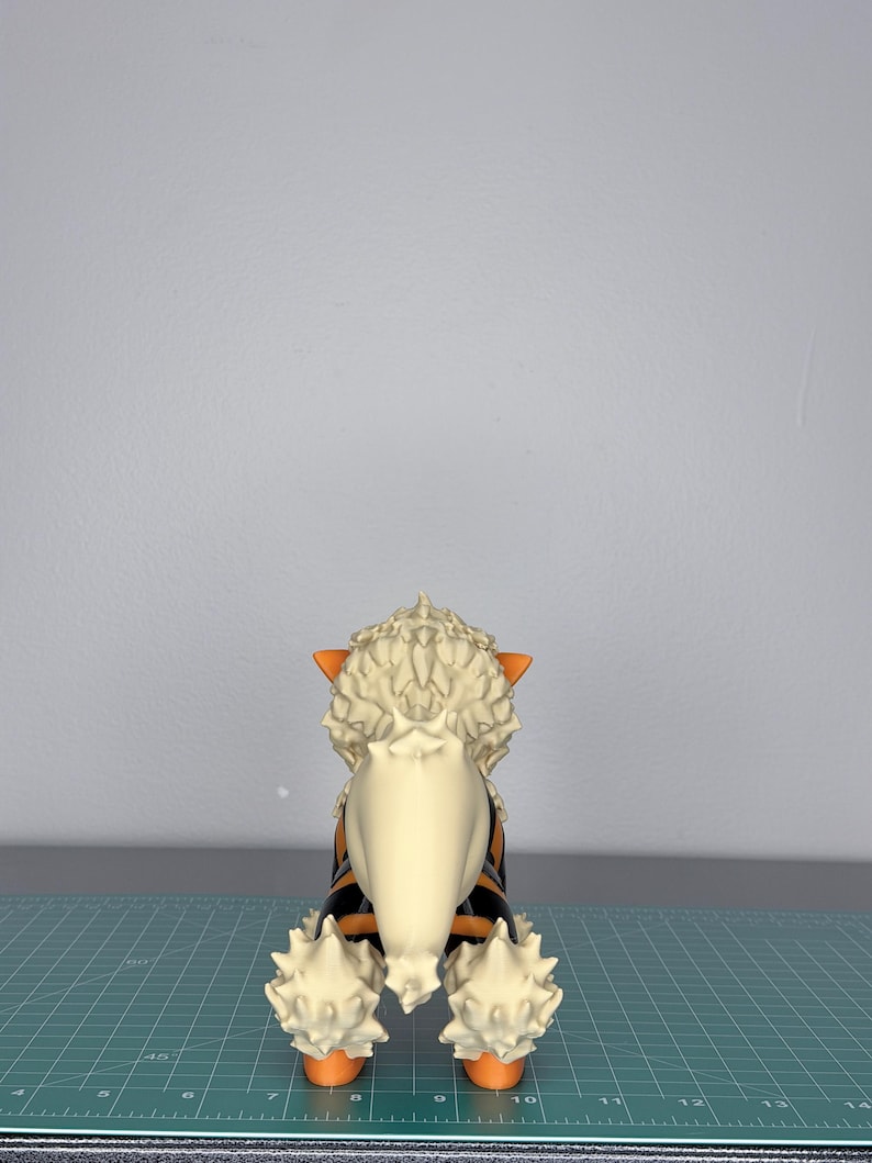 May include: A detailed figurine of a canine creature, viewed from the rear. The figure features a cream-colored mane and tail, with orange and black stripes on its body. The paws and ears are orange. The figurine is set against a gray background.