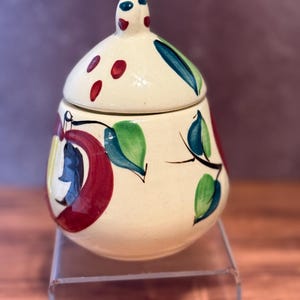 Może przedstawiać: Kremowy ceramiczny słoik z pasującą pokrywką, ozdobiony ręcznie malowanymi motywami czerwonych jabłek, zielonymi liśćmi i małymi czerwonymi kropkami. Słoik ma zaokrąglony kształt i mały uchwyt na pokrywce z niebieskimi i czerwonymi akcentami.