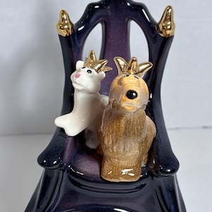 May include: A ceramic figurine featuring a white dog and a brown dog, both wearing gold crowns, sitting on a dark purple throne with gold accents. The throne has a high back and four legs, with a glossy finish on all surfaces of the piece.