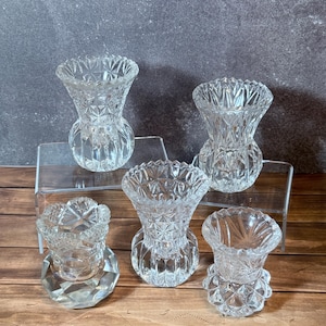 Può includere: Una collezione di cinque vasi in vetro trasparente con vari motivi geometrici complessi, inclusi disegni a diamante, a raggiera e a coste verticali. Ogni vaso ha una forma distinta, che spazia da basi bulbose a bordi sfaccettati.