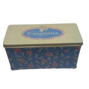May include: A rectangular metal storage tin with a cream-colored lid featuring a blue oval frame and the word Coupons in script. The sides of the tin are blue with a repeating pattern of small red flowers and green leaves throughout the design.