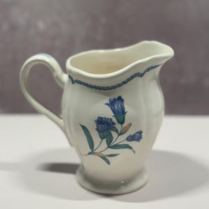 May include: A cream-colored ceramic creamer pitcher featuring a blue floral design on the front and a blue rope-style trim along the rim. The pitcher has a curved handle, a flared spout, and a rounded base with a smooth, glossy finish.