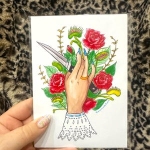 May include: A rectangular art print featuring a hand with blue fingernails holding a silver knife, surrounded by red roses and green Venus flytrap plants. The hand wears a gold ring and a white lace-trimmed sleeve with blue accents.