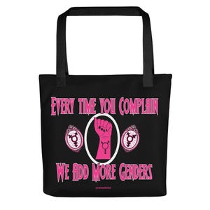 May include: A black tote bag with black handles featuring a pink graphic of a raised fist with a gender symbol on the wrist, flanked by two ornate oval frames containing gender symbols, and the text Every Time You Complain We Add More Genders.
