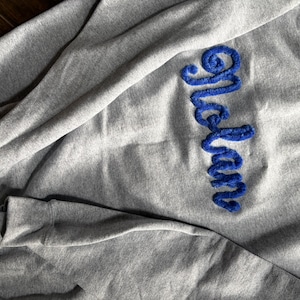 K&ouml;nnte beinhalten: Ein hellgraues Sweatshirt mit Rundhalsausschnitt, auf dessen Brust der Name Nolan in einer leuchtend blauen, strukturierten und flauschigen Schreibschrift eingestickt ist. Das Kleidungsst&uuml;ck hat lange &Auml;rmel und einen gerippten Halsausschnitt.