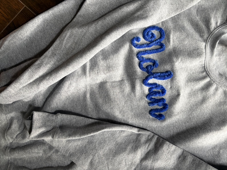 K&ouml;nnte beinhalten: Ein hellgraues Sweatshirt mit Rundhalsausschnitt, auf dessen Brust der Name Nolan in einer leuchtend blauen, strukturierten und flauschigen Schreibschrift eingestickt ist. Das Kleidungsst&uuml;ck hat lange &Auml;rmel und einen gerippten Halsausschnitt.