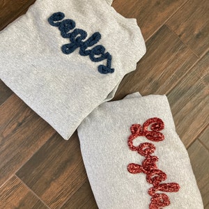 K&ouml;nnte beinhalten: Zwei hellgraue Sweatshirts mit Rundhalsausschnitt und strukturierten Chenille-Schriftz&uuml;gen. Ein Sweatshirt zeigt das Wort "eagles" in Dunkelmarineblau, w&auml;hrend das andere das Wort "Loyola" in tiefem Bordeauxrot in Schreibschrift zeigt.