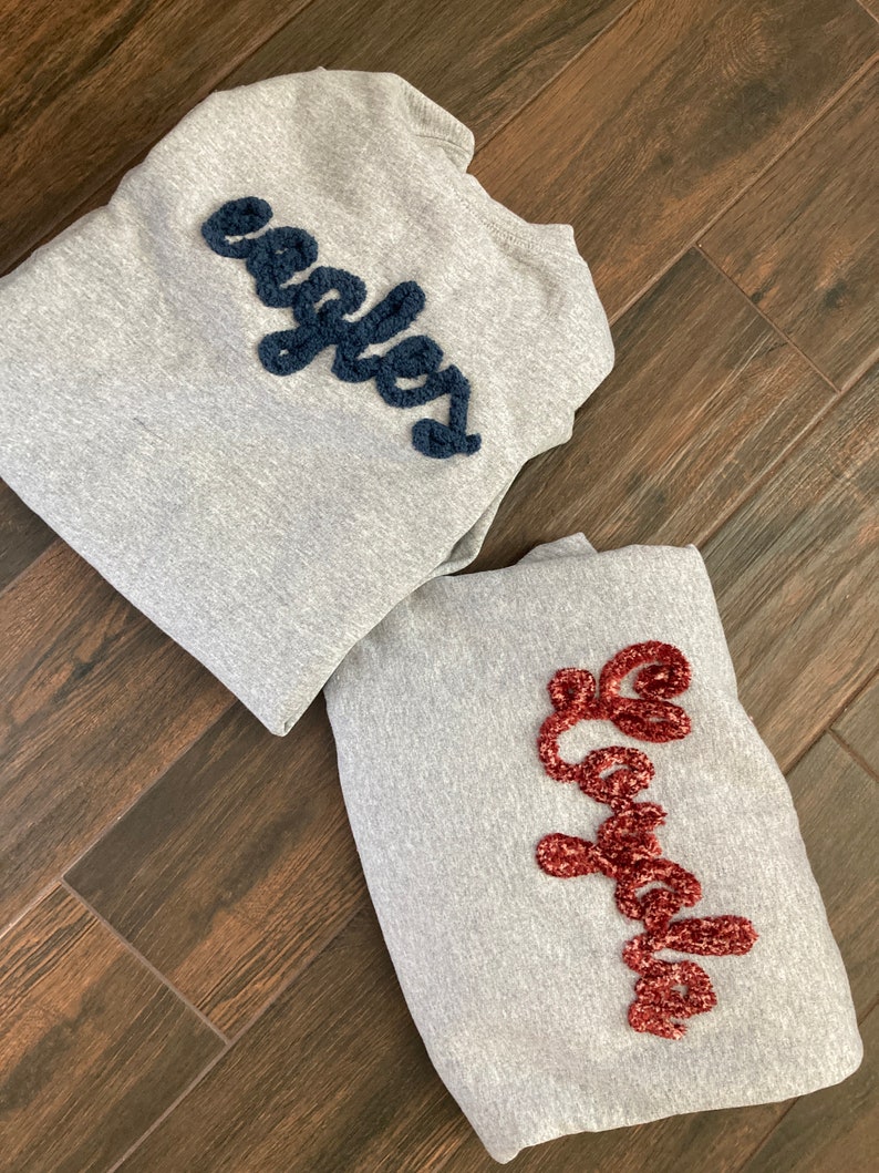 K&ouml;nnte beinhalten: Zwei hellgraue Sweatshirts mit Rundhalsausschnitt und strukturierten Chenille-Schriftz&uuml;gen. Ein Sweatshirt zeigt das Wort "eagles" in Dunkelmarineblau, w&auml;hrend das andere das Wort "Loyola" in tiefem Bordeauxrot in Schreibschrift zeigt.