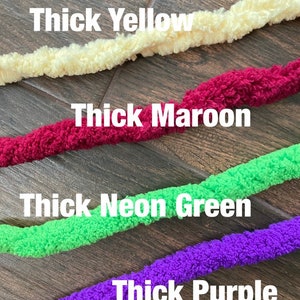 K&ouml;nnte beinhalten: Vier Str&auml;nge aus dickem, strukturiertem Garn in Gelb, Kastanienbraun, Neongr&uuml;n und Lila. Die Textbeschriftungen Thick Yellow, Thick Maroon, Thick Neon Green und Thick Purple befinden sich &uuml;ber jedem entsprechenden Garnstrang im Bild.
